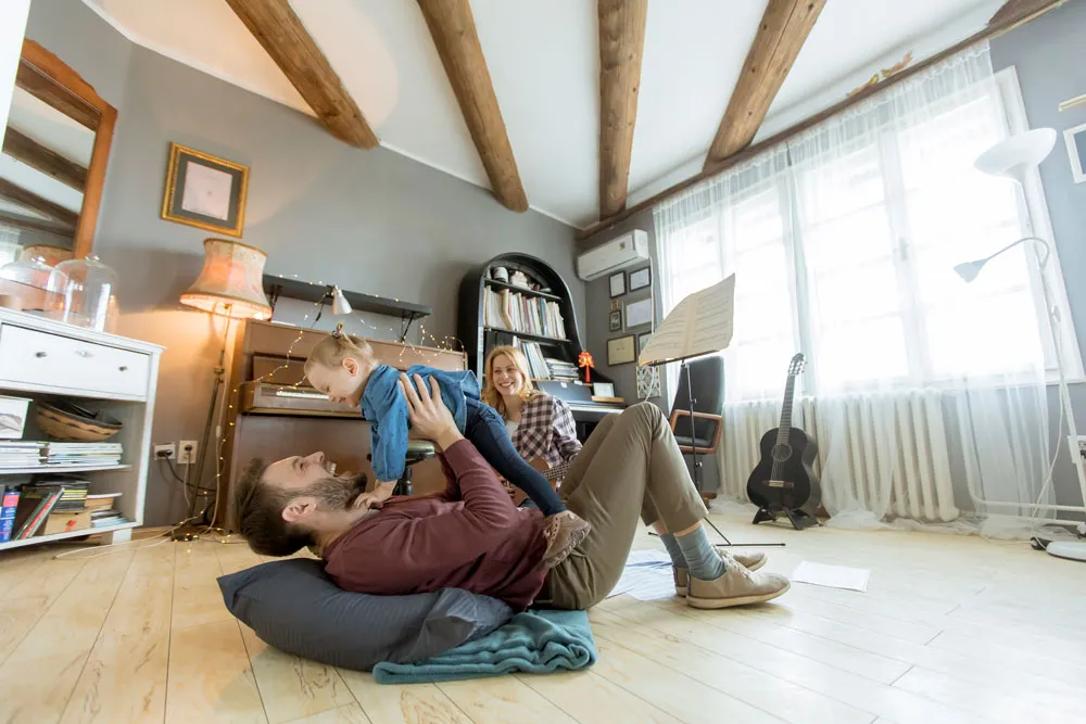 Family enjoying time together indoors, with a father playfully lifting a child, while a mother smiles in a cozy, well-decorated living room featuring wooden beams and natural light.