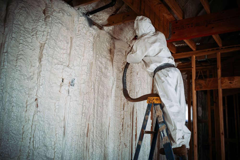 Worker applying spray foam insulation to walls in a construction setting, wearing protective gear, demonstrating energy-efficient insulation solutions for buildings in Western Australia.